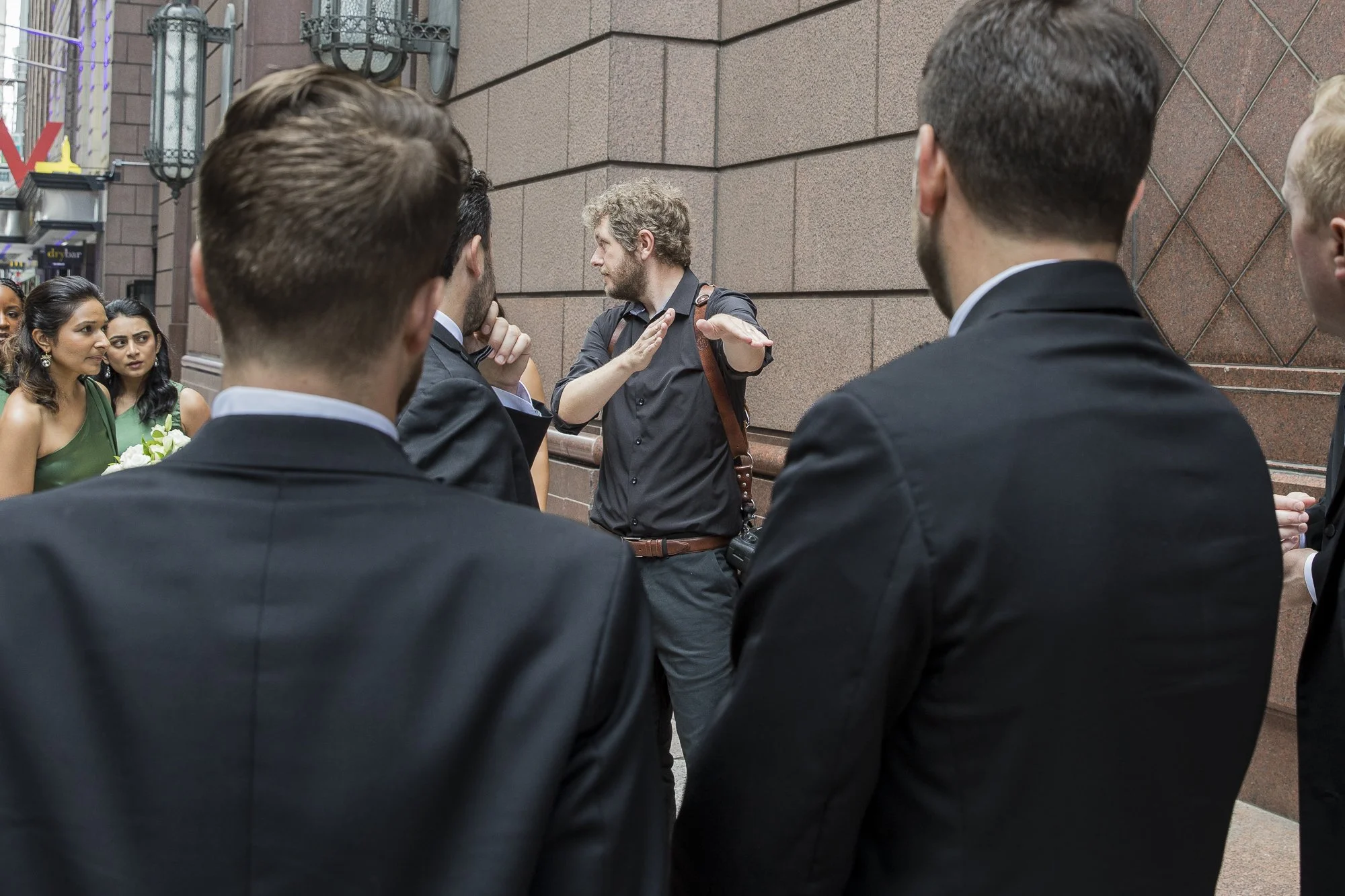 A man with curly hair and a beard is gesturing with his hand while talking to a group of people dressed in formal attire outside, next to a building with a brick wall.