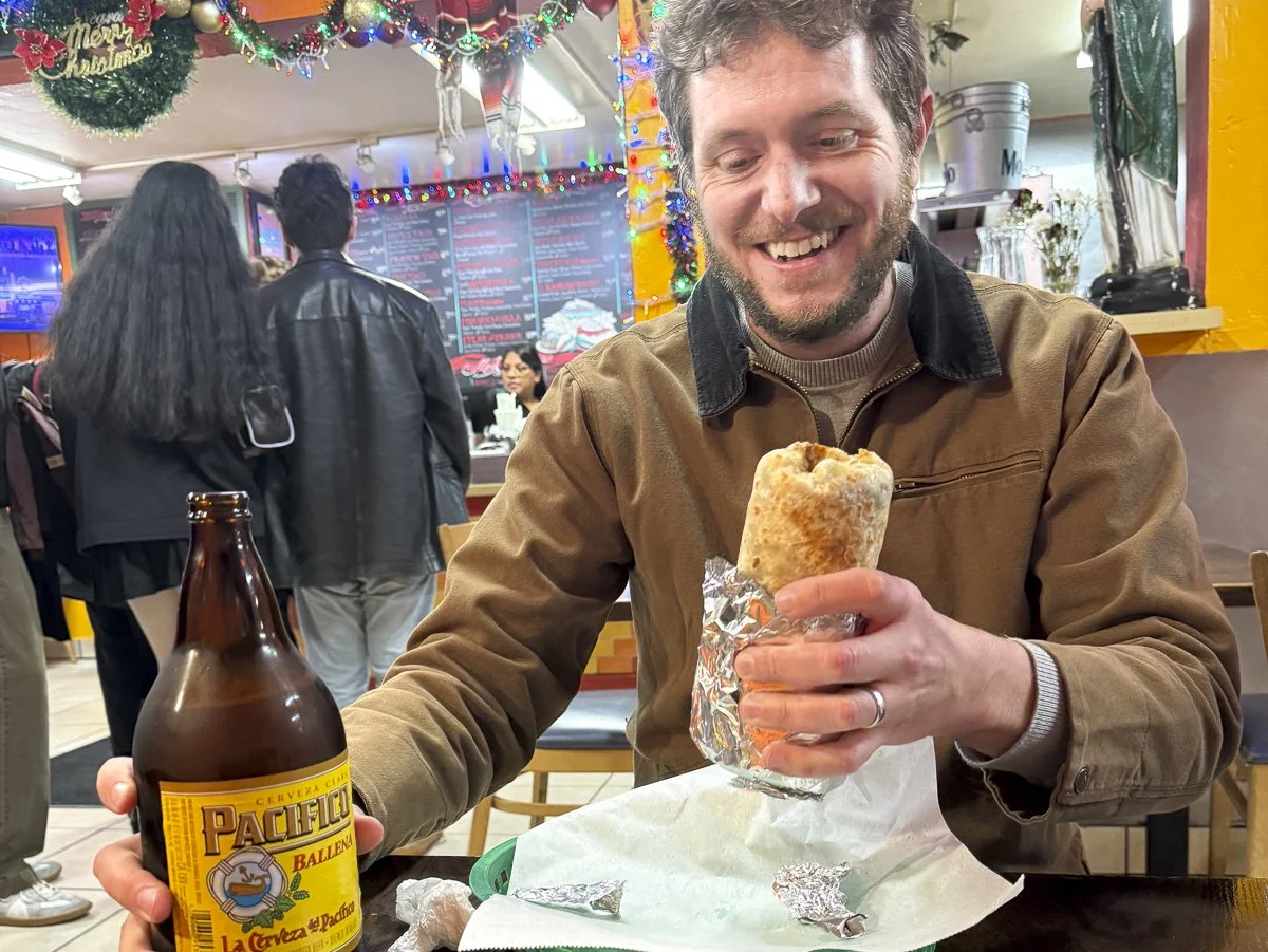 A smiling man with a beard and brown jacket sitting at a restaurant table holding a large burrito wrapped in foil in one hand and a large bottle of Pacifico beer in the other. The restaurant is decorated with Christmas lights and ornaments, and a Christmas wreath with a