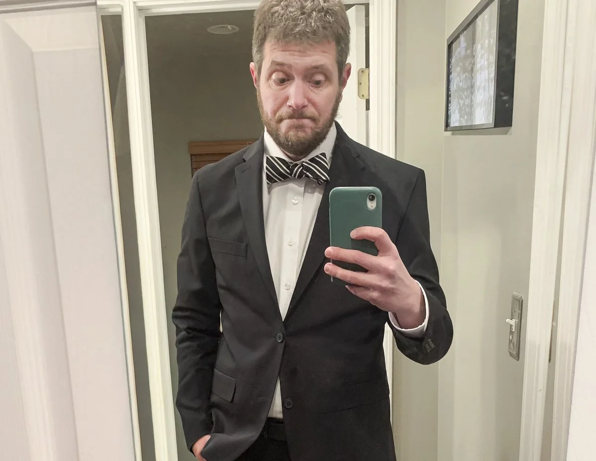A man in a black tuxedo with a bow tie taking a selfie in a mirror.