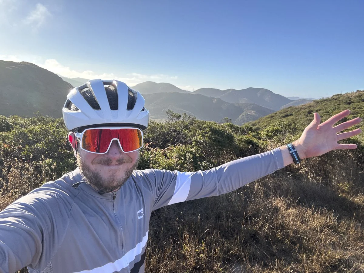 A man wearing a white biking helmet and red reflective sunglasses taking a selfie with outstretched arm in a mountainous outdoor setting.