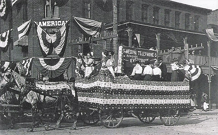 The Medina Telephone Company fancy parade wagon participates in the village 4th of July Independence Day festivities in 1912 as it passes by the decorated American House Hotel owned by Warren J. Anderson on the corner of East Liberty and North Court Streets.