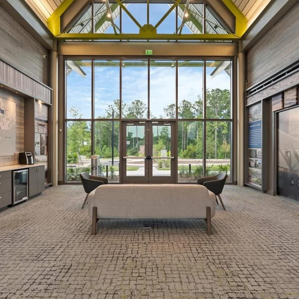 Interior of a modern building with large glass windows and doors, a beige sofa, two black armchairs, and a kitchenette area on the left, with trees visible outside.