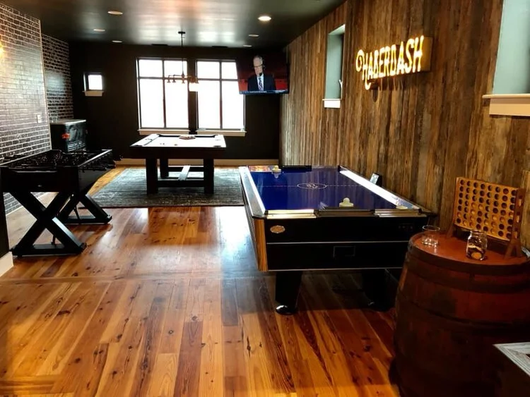 Indoor game room with shuffleboard and foosball tables, a pool table, a mounted TV, and a sign that reads 'QUIDASH' on wooden wall, with hardwood flooring and a window with natural light.