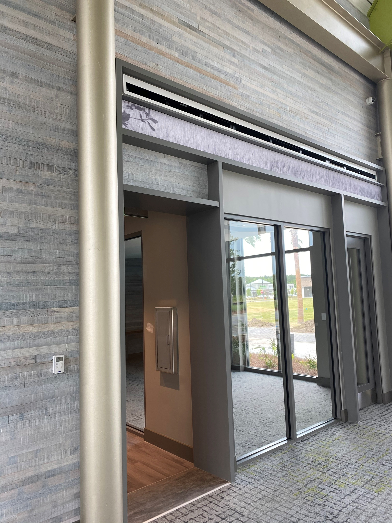 Modern wind shelter with glass walls and sliding door in an indoor space with textured wall and carpeted floor.