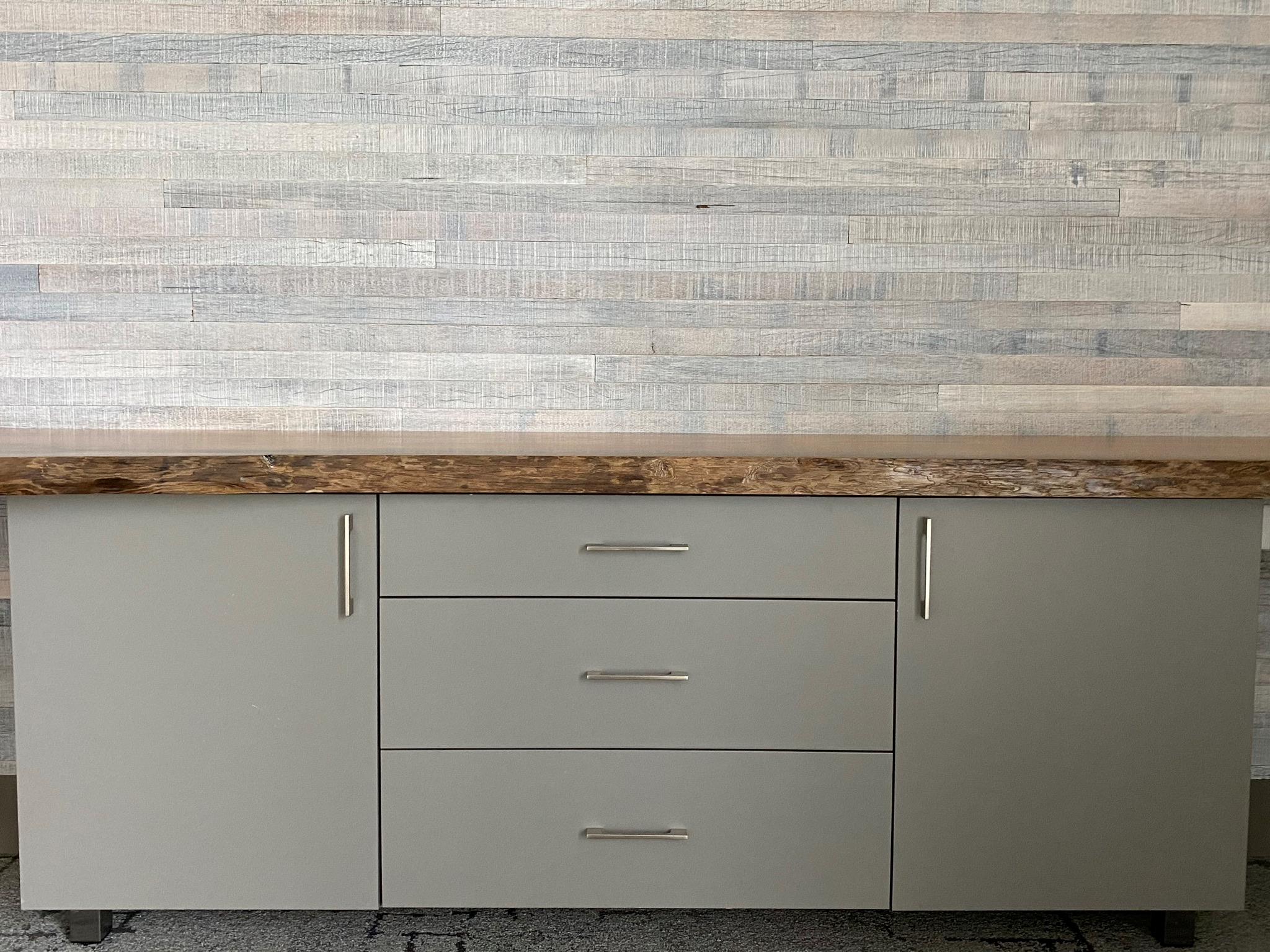 A modern kitchen cabinet with a dark natural wood countertop, gray doors, and drawers with horizontal silver handles. The cabinet is placed against a light-colored textured wall.