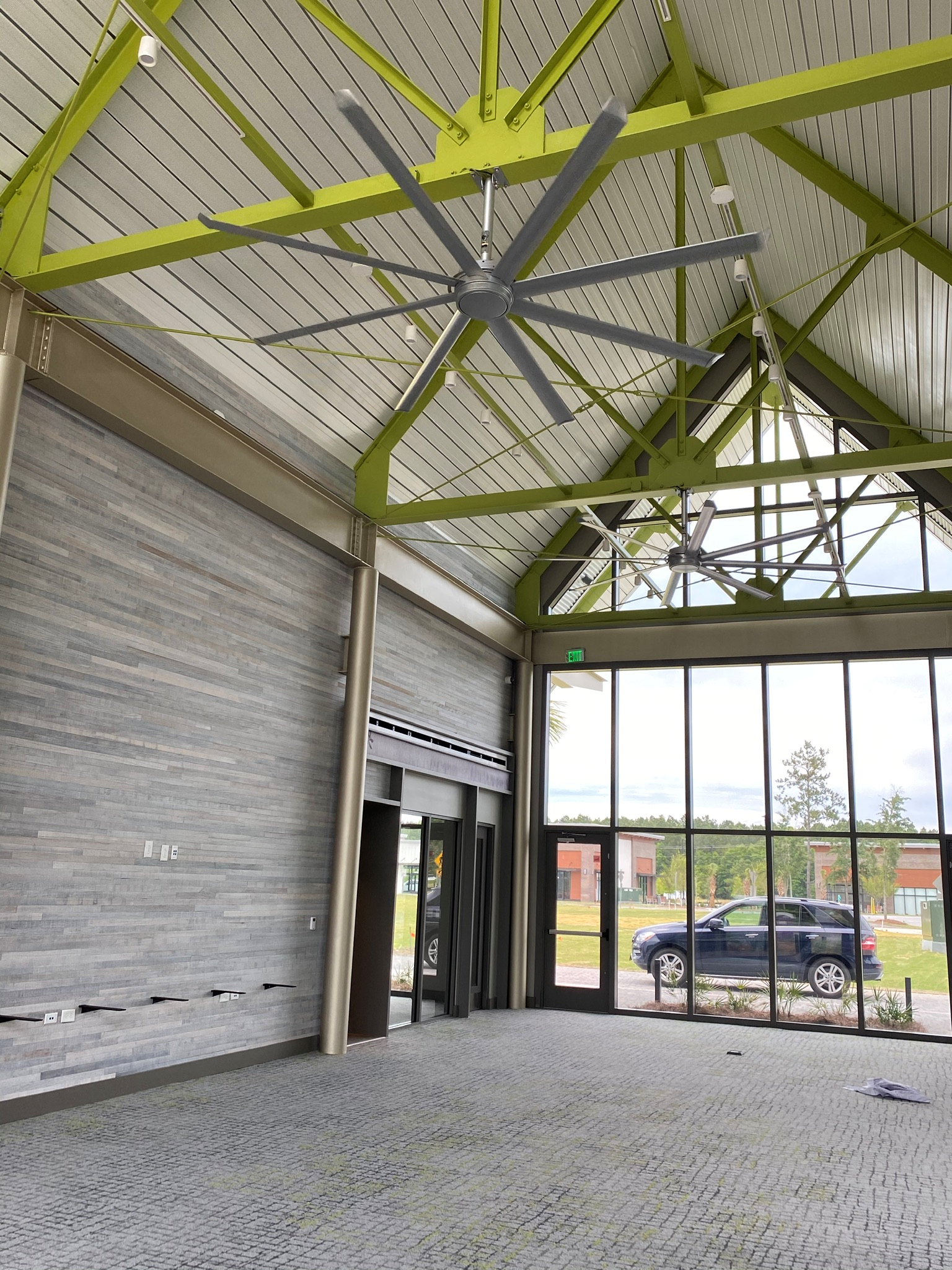 Interior of a modern building with a high ceiling, large glass windows, and ceiling fans, overlooking a parking lot with a black car and some trees.