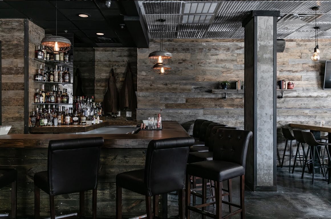 Interior of a modern bar featuring wooden walls, a wooden bar counter with black barstools, shelves stocked with various alcohol bottles, and hanging pendant lights with Edison bulbs.