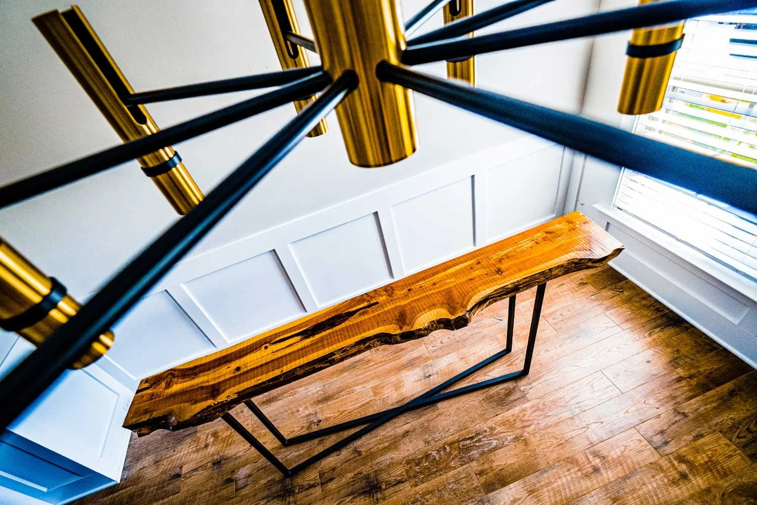 A wooden table with a natural, live edge and black metal legs in a room with hardwood floors and white paneled wainscoting, viewed from above near a window with blinds.