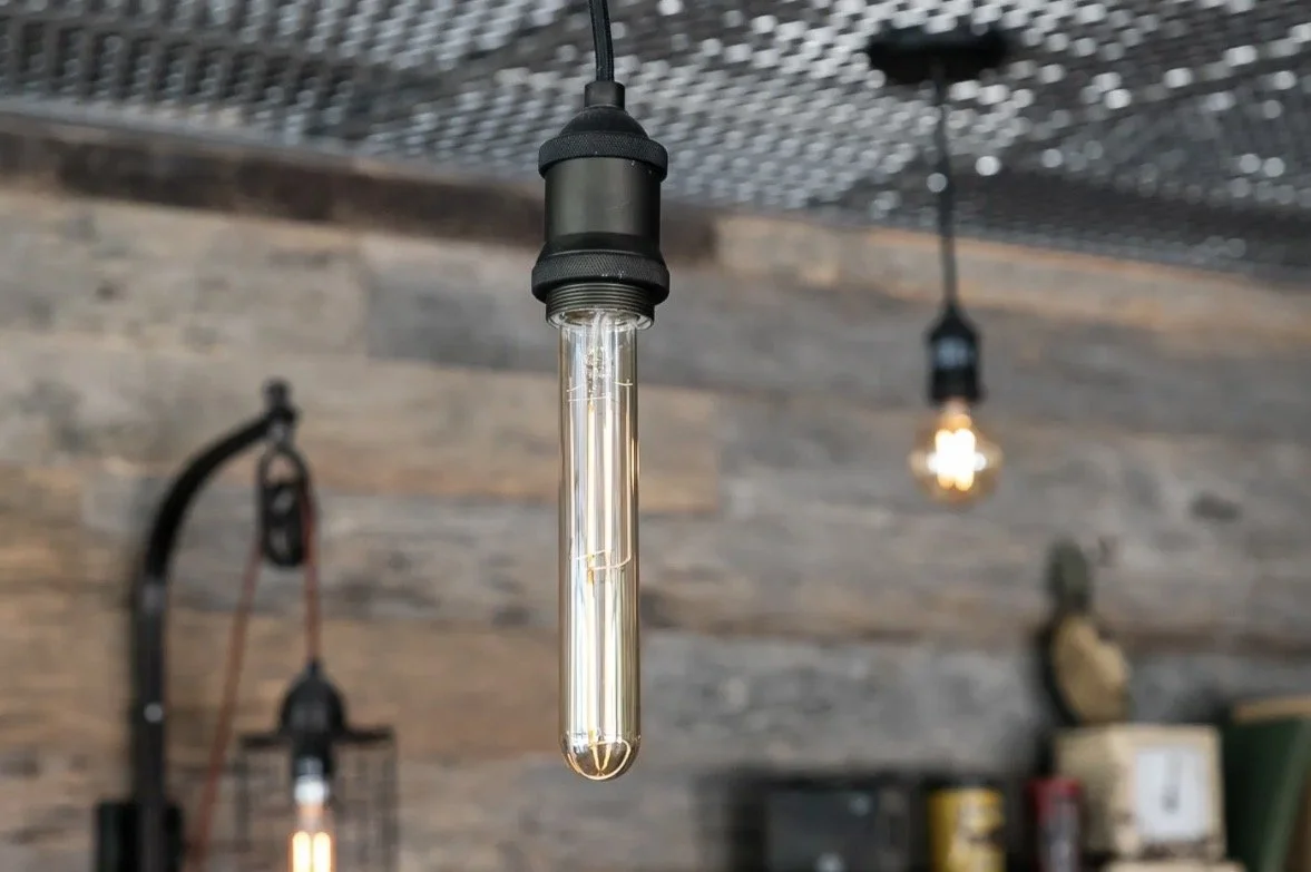 Close-up of a vintage-style Edison light bulb hanging from a black cord in a rustic interior with wooden wall panels and other exposed bulbs in the background.