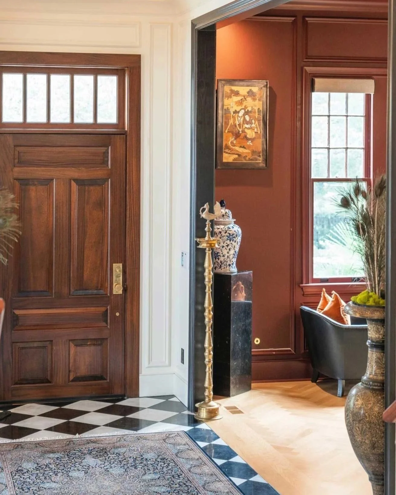 Interior view of a home entrance with a wooden front door, checkered black and white floor, and a glimpse into a sitting area with red walls, a framed painting, large window, black armchair, and decorative vases.