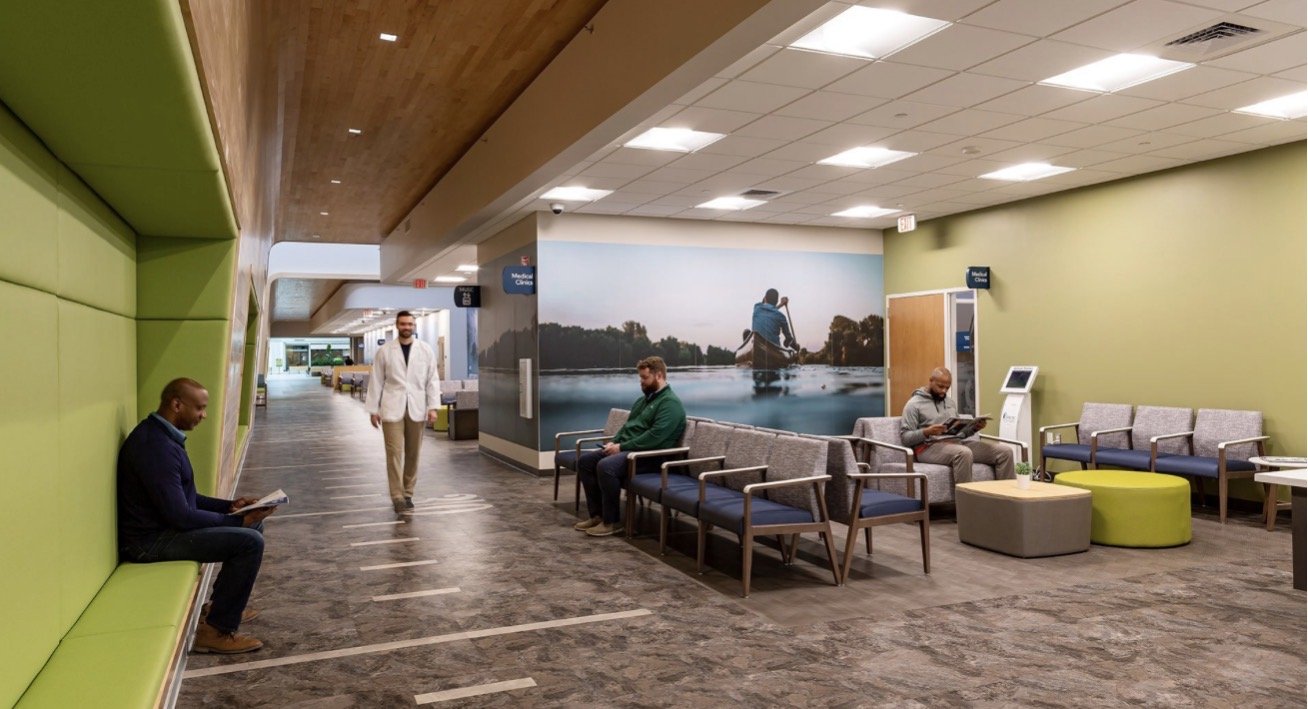 Hospital waiting area with chairs, digital kiosk, medical clinic sign, large wall mural of a person kayaking on a river, and several people sitting and reading.