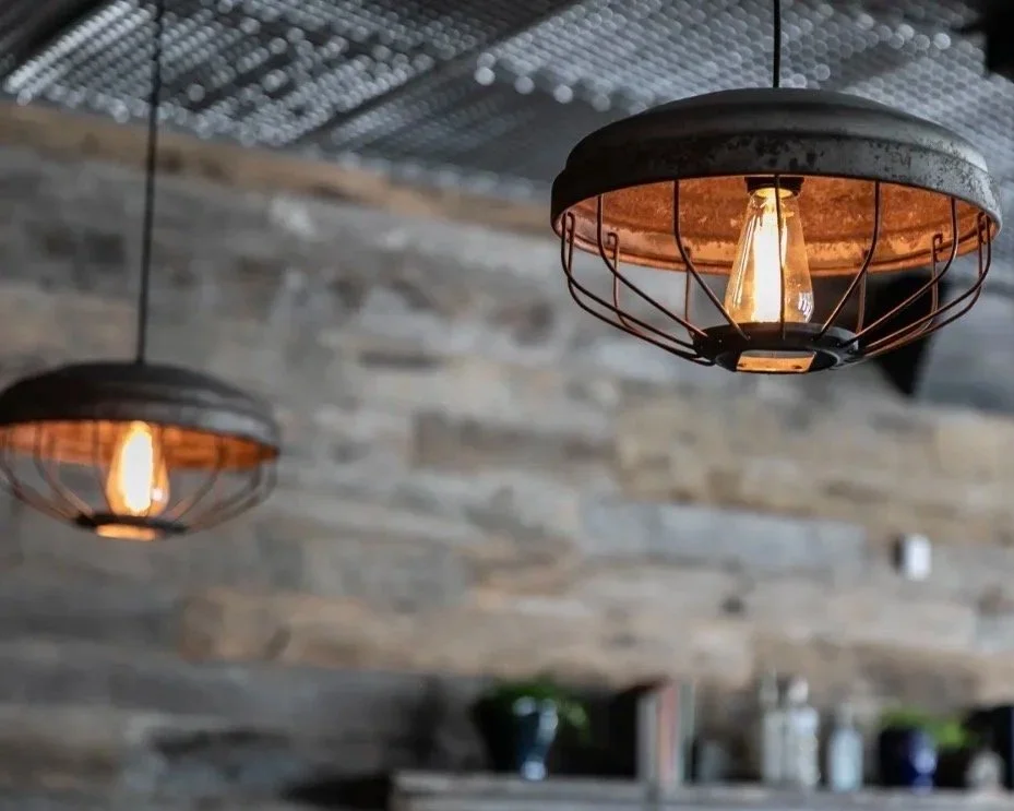 Close-up of two industrial-style hanging ceiling lamps with exposed Edison bulbs, in front of a rustic stone wall.