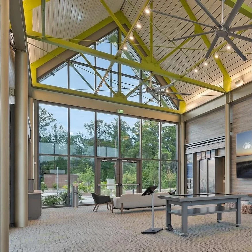 Indoor lobby area with high ceiling, large glass windows, modern furniture, and ceiling fan, illuminated by natural light.