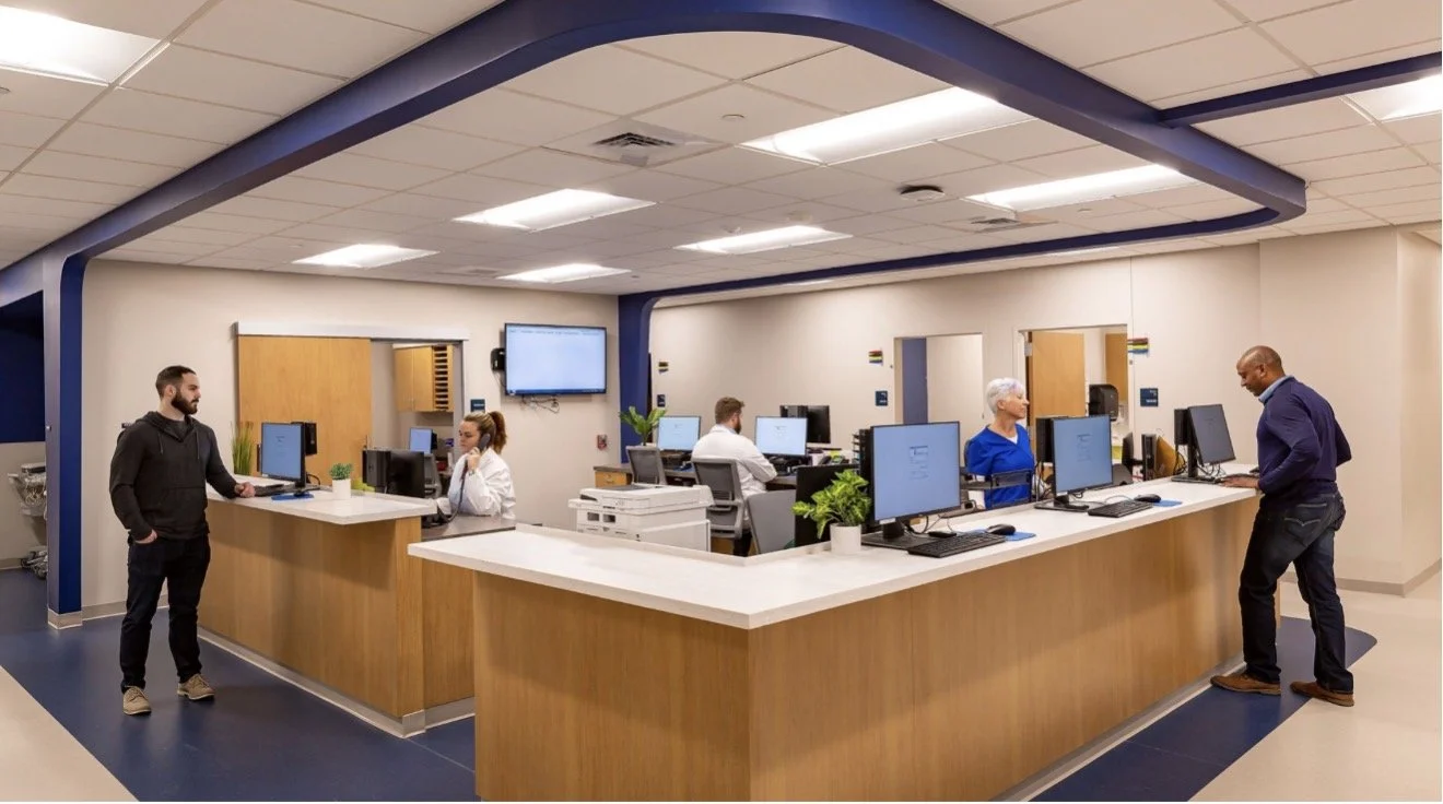 Hospital reception area with staff and patients, multiple computer workstations, and a wooden front desk.