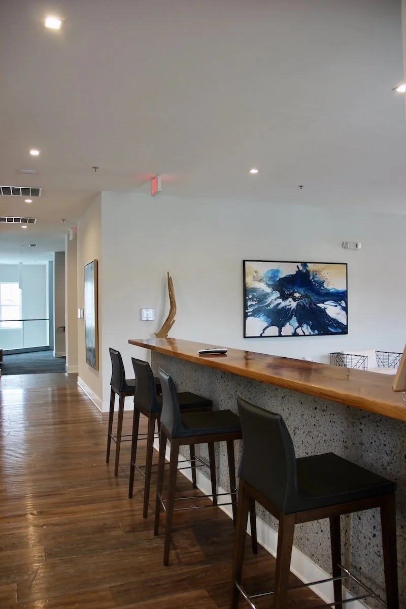 A modern indoor interior with a wooden bar counter and five bar stools with black seats. There is a piece of decorative driftwood on the counter, and a blue abstract painting on the white wall behind it. The space has wooden flooring and ceiling lights.