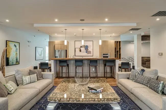 Modern living room with light-colored sofas, a marble coffee table, and a kitchen with a breakfast bar behind. Art pieces hang on the walls, and there are decorative pillows and a rug in the space.