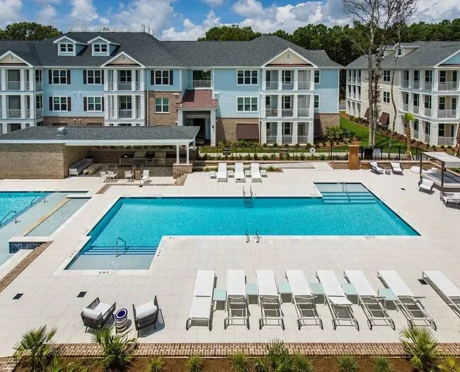 Outdoor swimming pool area in front of a multi-story apartment complex with poolside lounge chairs and a covered seating area, surrounded by landscaped greenery and trees.
