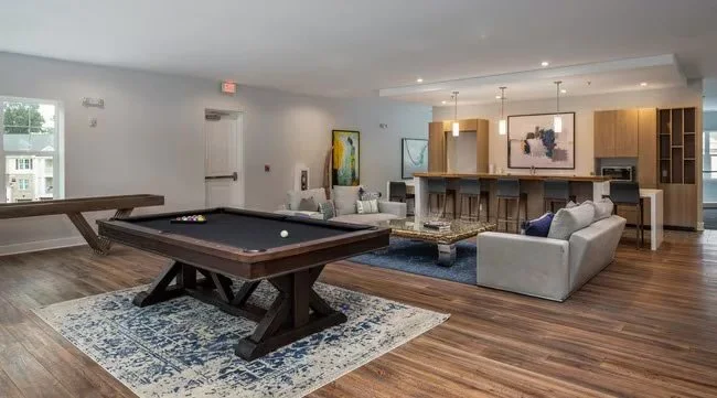 A spacious, modern living room with a pool table on a decorative rug, seating area with sofas and chairs, open kitchen with bar stools, and hardwood floors.