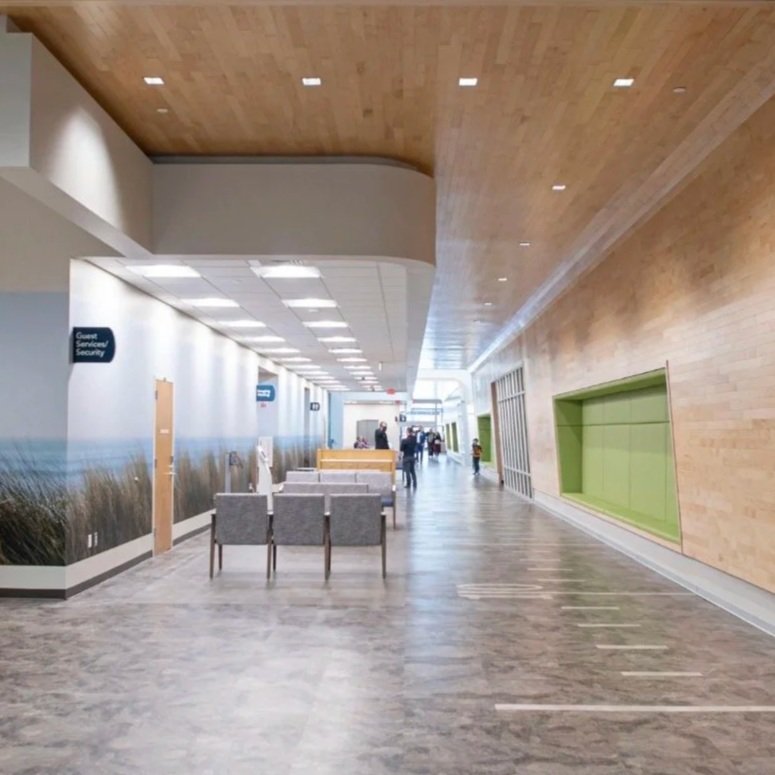 Indoor corridor with wooden ceiling, white walls with nature mural, gray tiled floor, green wall niches, seating area, and a few people walking in the distance.