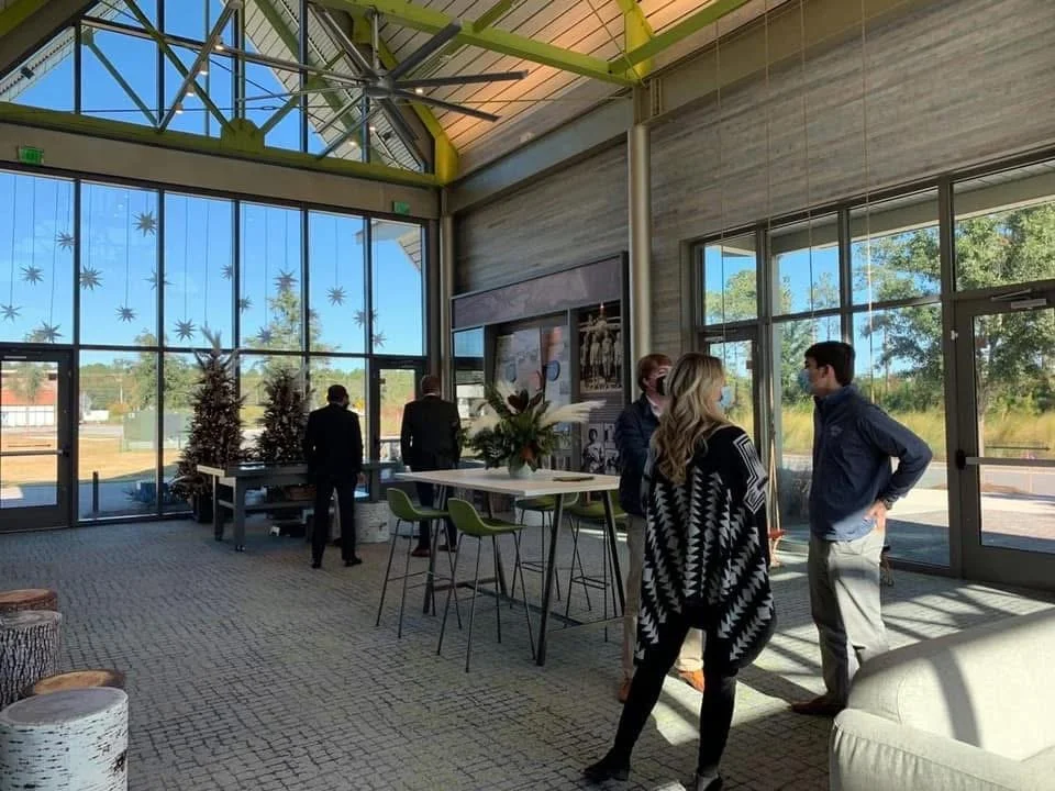 People inside a modern, glass-walled building with natural sunlight, standing and conversing near a tall table with chairs, decorated with plants, and Christmas trees in the background.