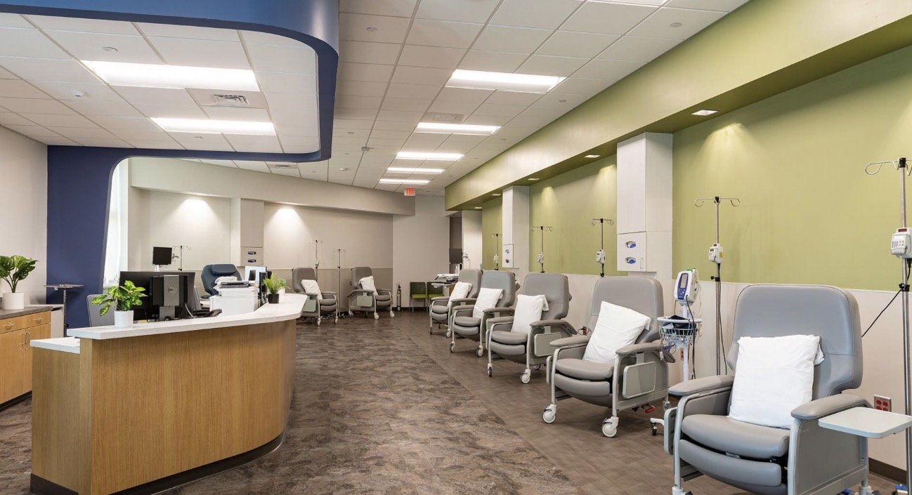 A row of medical chairs with attached IV stands in a hospital or clinic room, with a reception desk in the foreground and a green wall in the background.