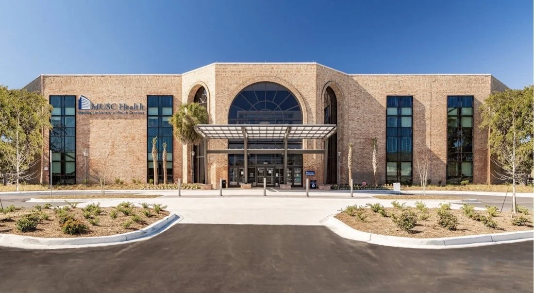 Front view of a modern brick hospital building with a large arched window and glass doors, surrounded by a parking lot and landscaped yard with small trees and bushes.