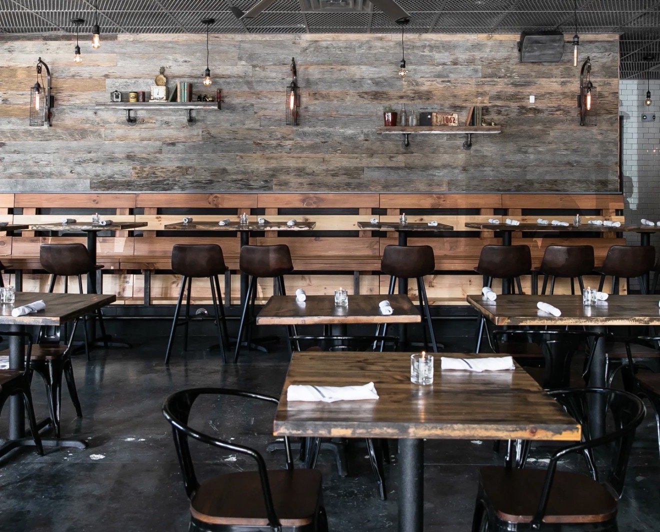 Interior of a modern restaurant with wooden tables, black chairs, and a textured wooden wall decorated with books and small decorative items, lit by hanging Edison bulbs.