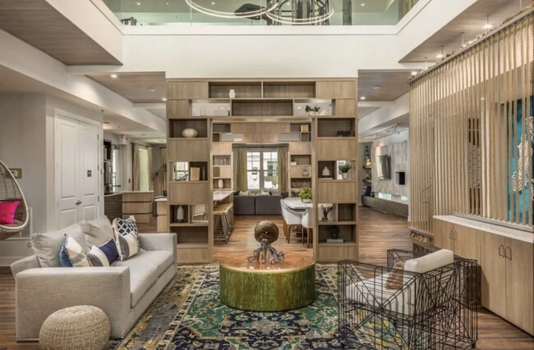 Modern living room with a white sofa, decorative pillows, a round ottoman, and an intricate coffee table, separated by a large wooden shelving unit, with a dining area and a pool table in the background, and natural light from windows and a skylight.