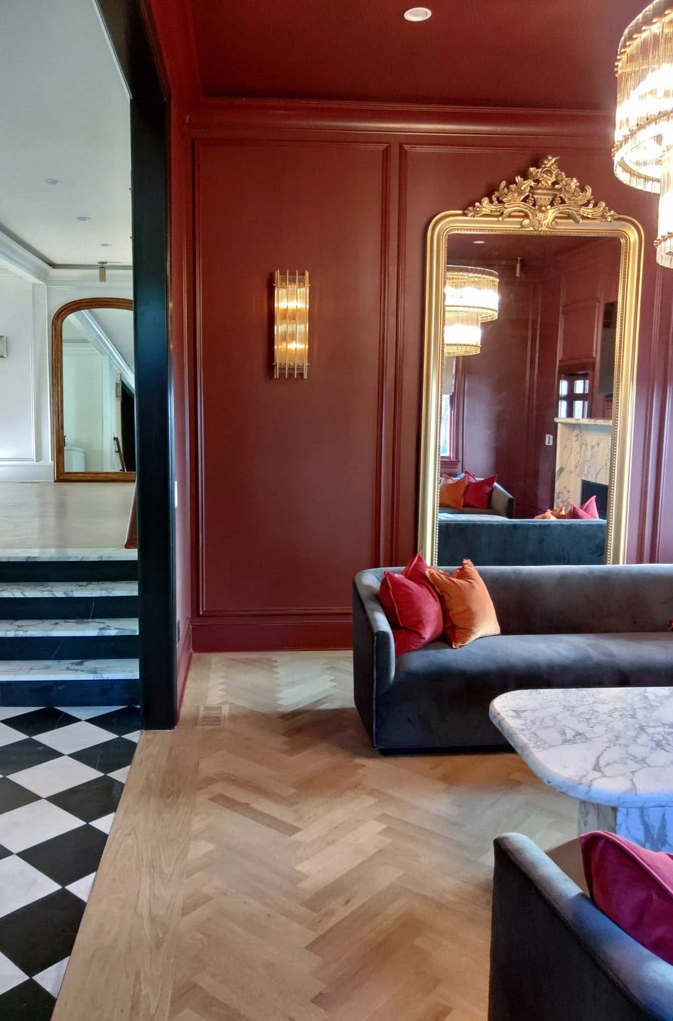 Living room with a black velvet sofa, colorful pillows, a marble coffee table, a large gilded mirror, a red wall with a gold wall sconce, and a chandelier.