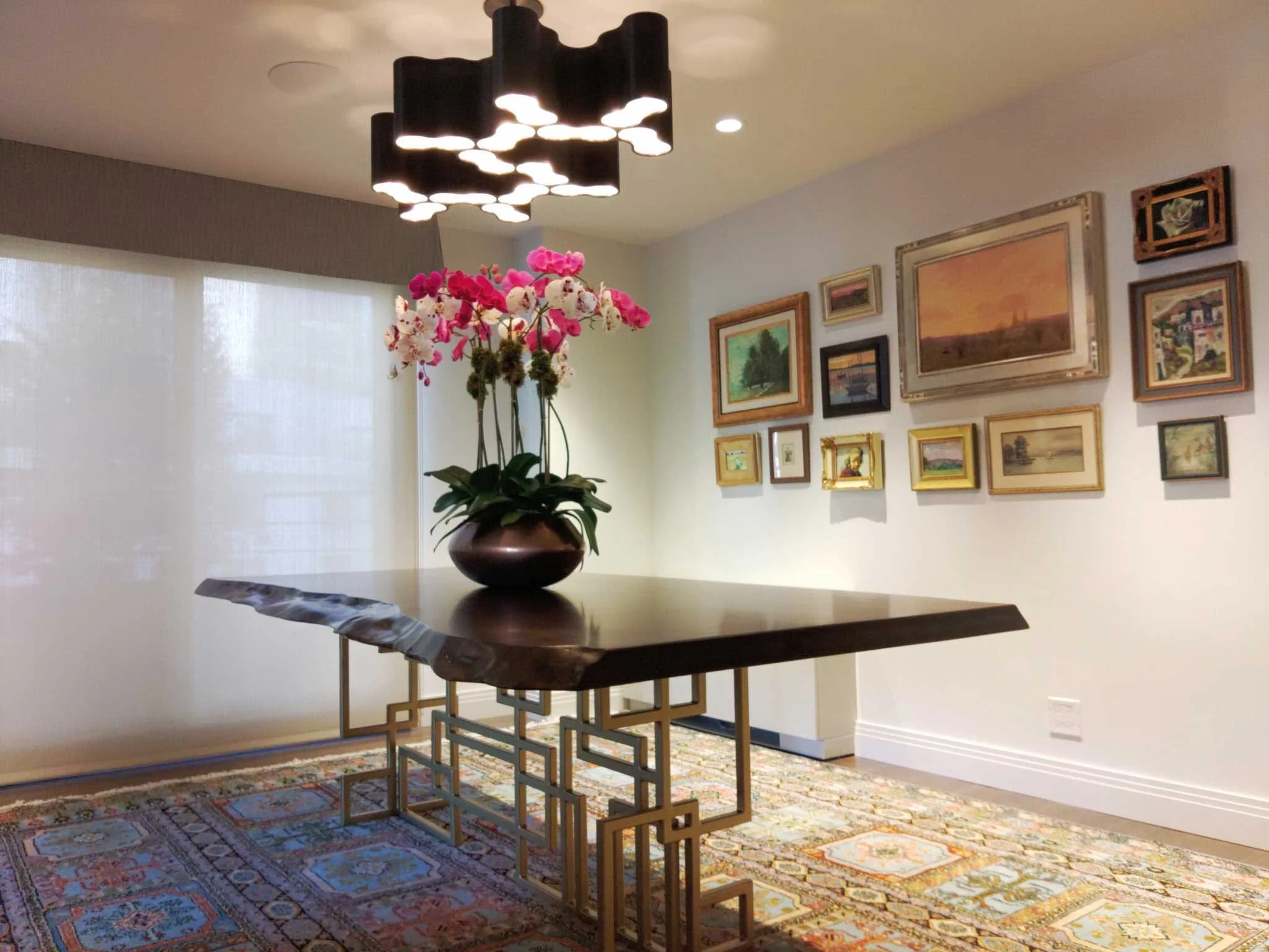 A modern living room featuring a wooden table with a decorative metal base, topped with a large vase holding pink and white orchids. The background has a gallery wall with various framed paintings and a large window with white curtains, and a patterned rug on the floor.