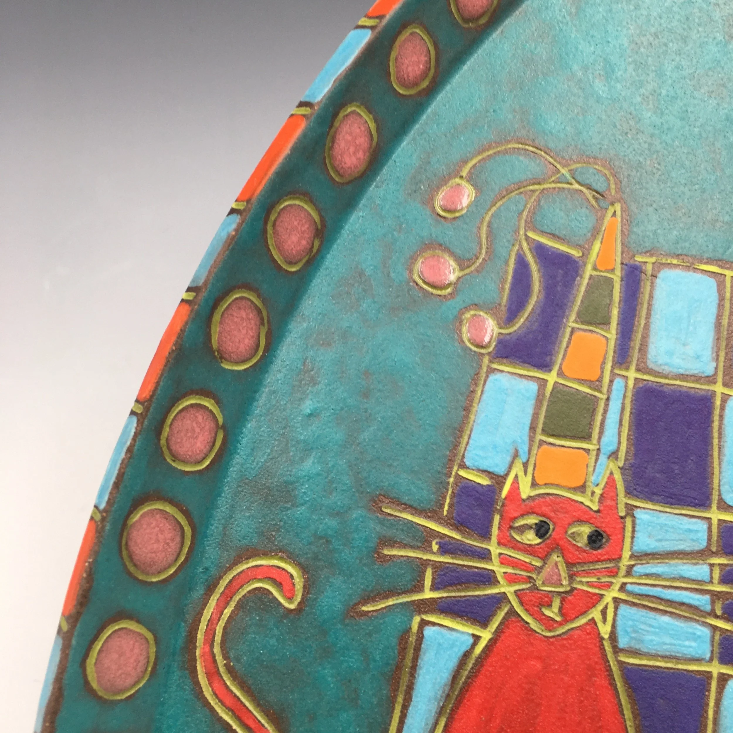 Triangular ceramic plate with a red cat wearing a hat sitting on a yellow and orange carpet in front of a blue and purple building next to a striped candle.