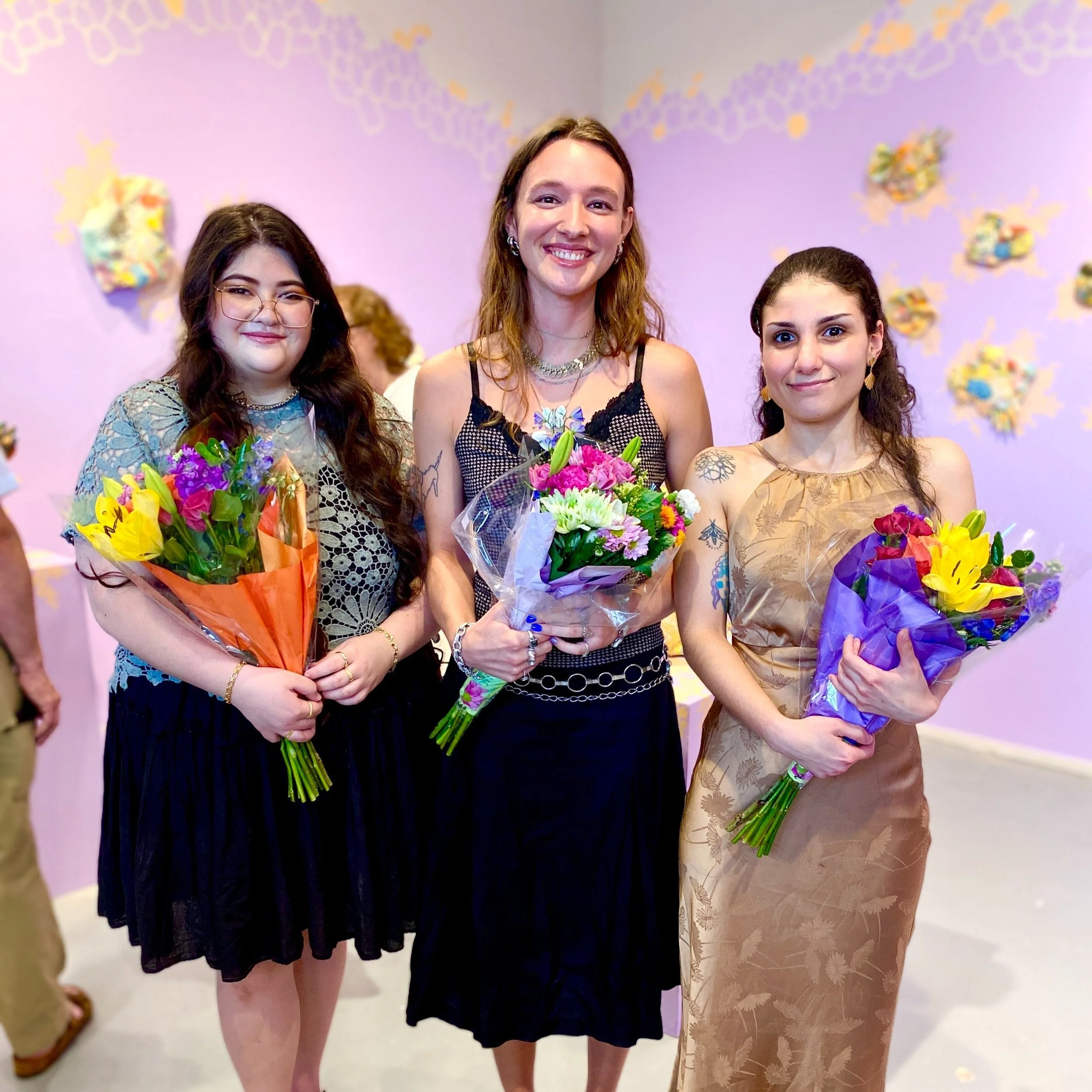 Resident Artists Christine, Carley, and Steph stand together in front of the exhibitions, holding bouquets of flowers.