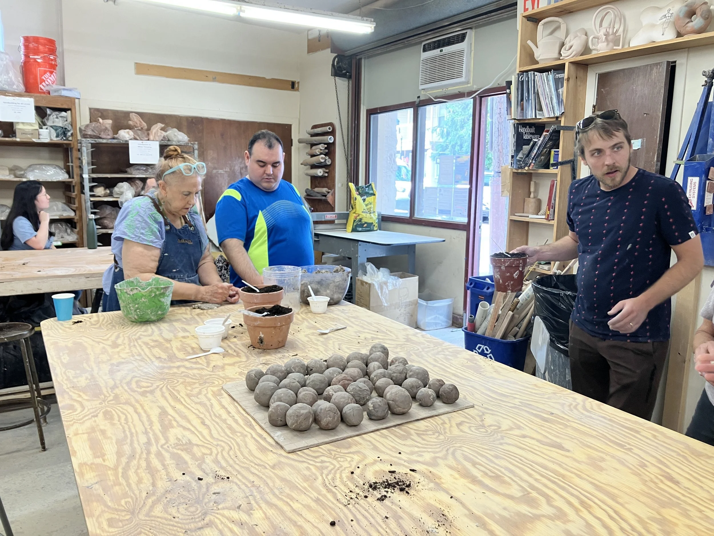 Three people gather around the wooden work bench making seed bombs. A large pile of finished seed bombs is in the middle of the table