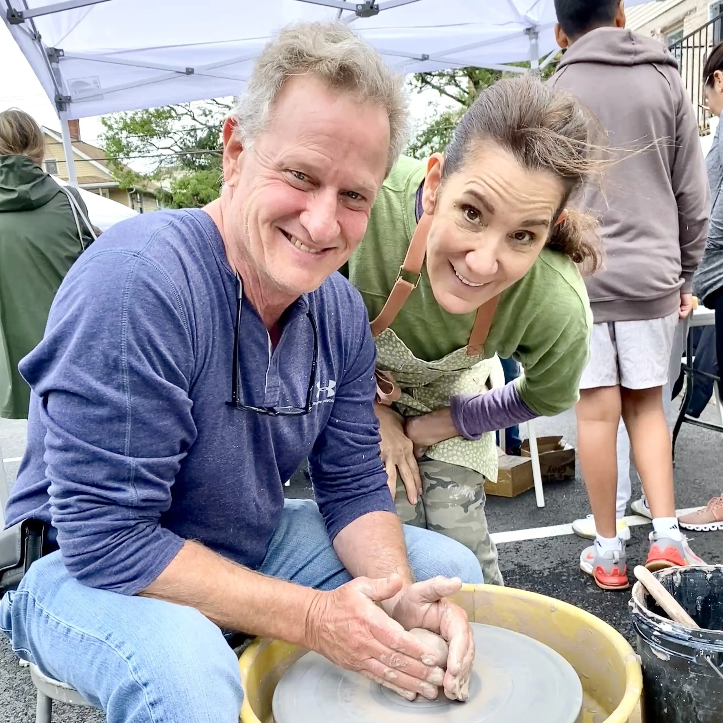 A man and a woman smile at the camera. The man is centering clay on a wheel.