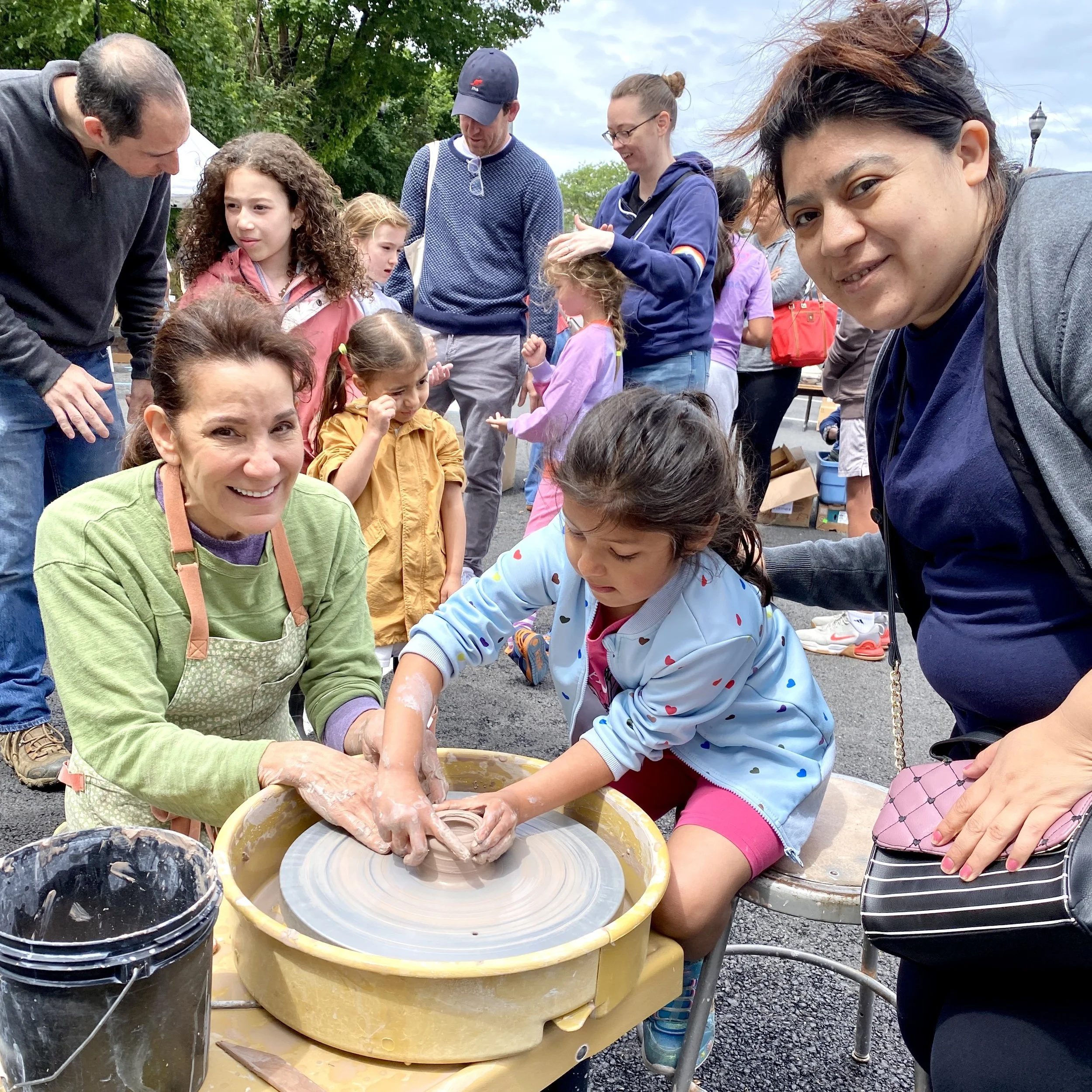 A woman helps a young girl center clay on the wheel while another woman looks on