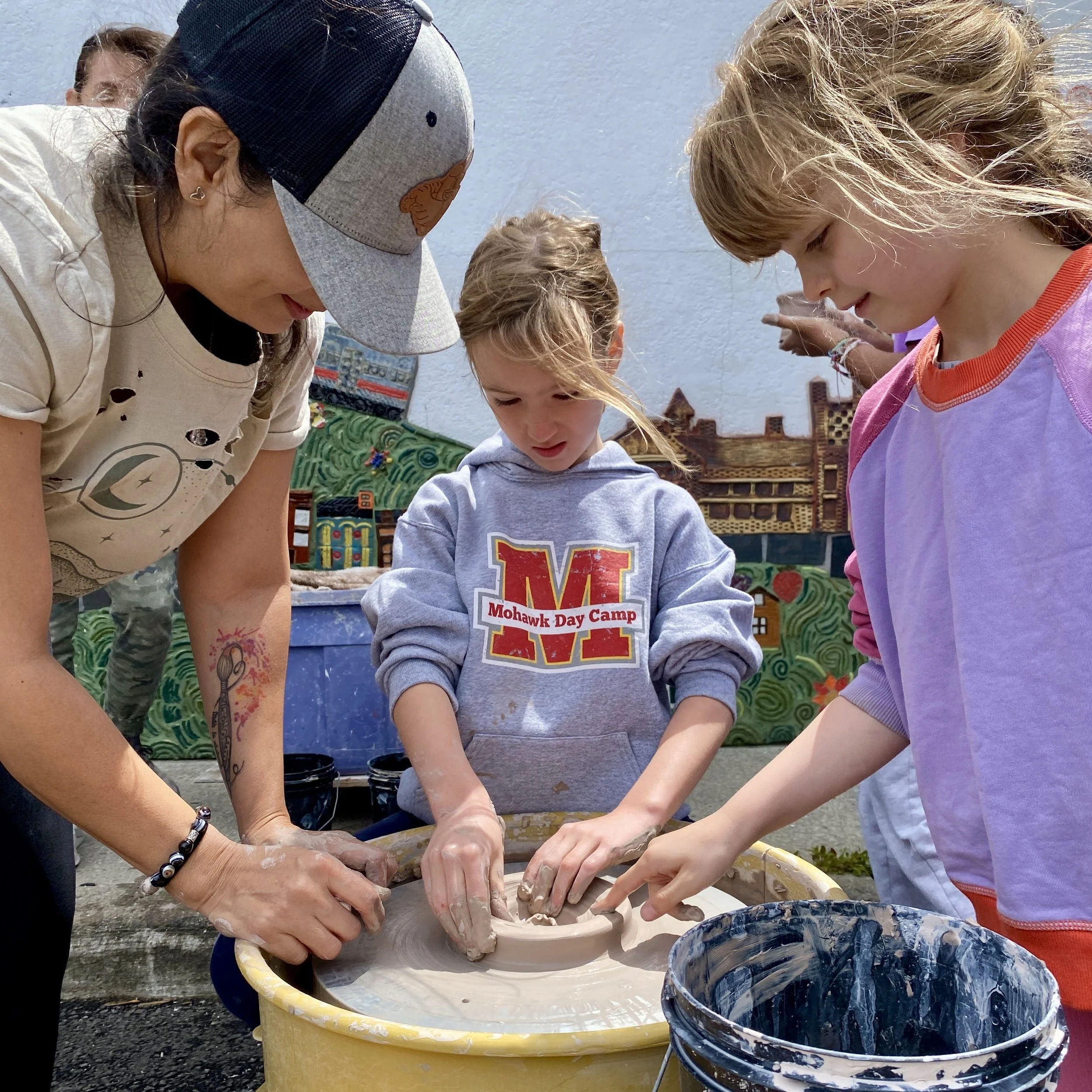 A woman helps a young girl center clay on a wheel, while another young girl reaches over to feel the clay