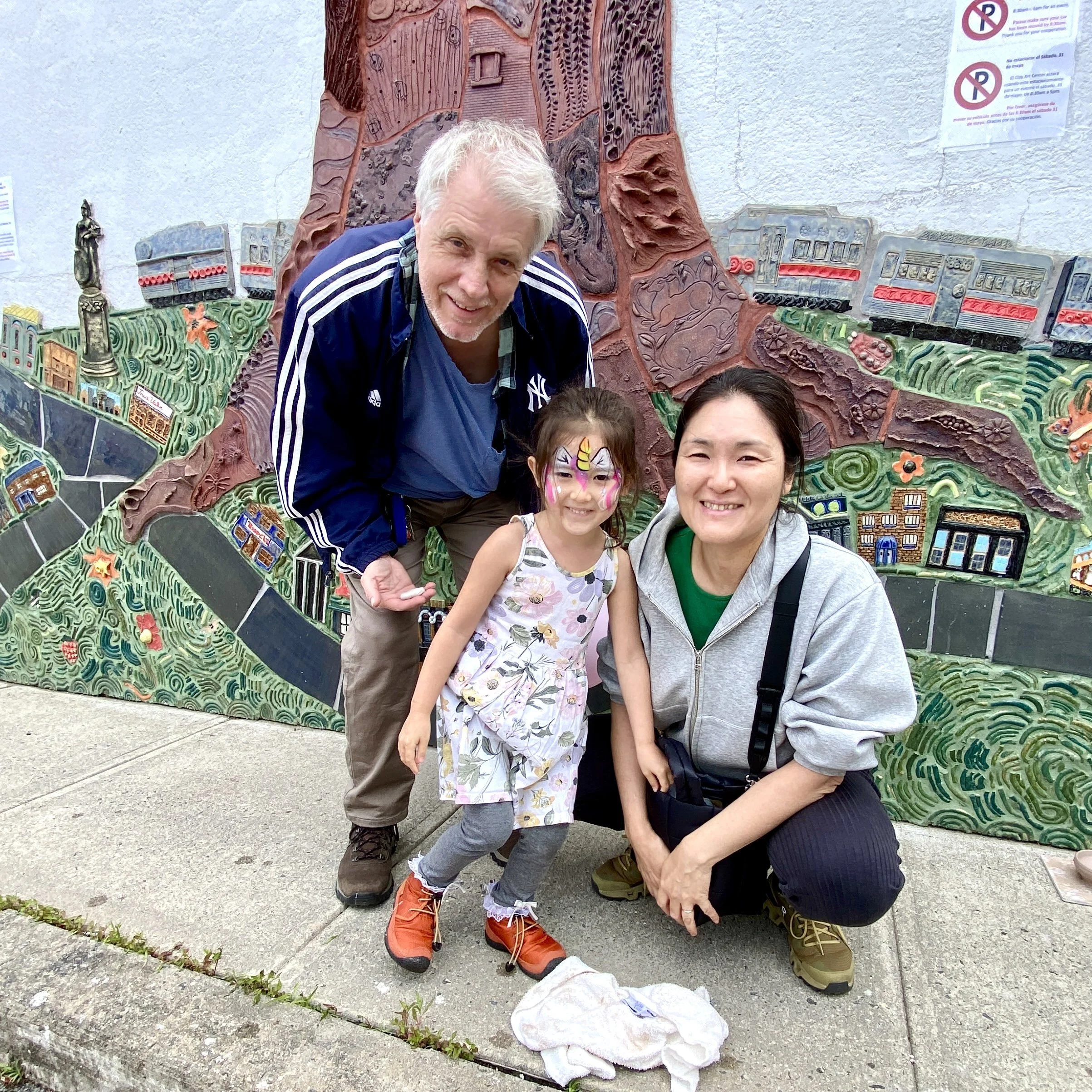 A young girl, man, and woman smile for a group photo. A ceramic mural of a tree is visible behind them