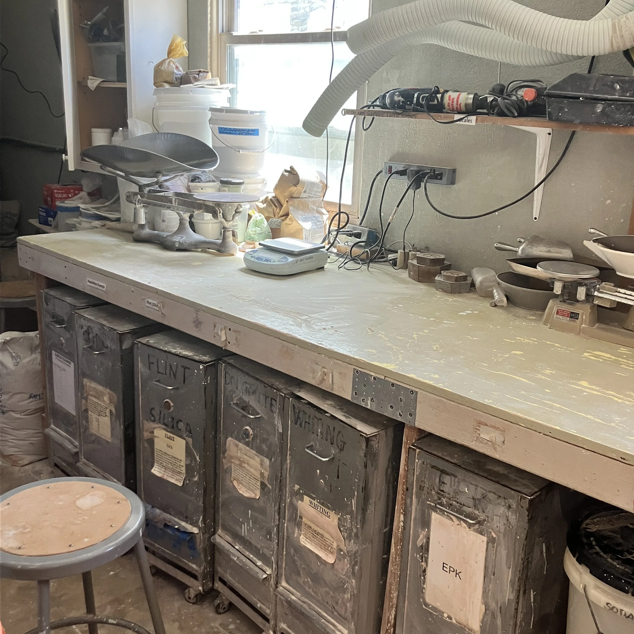 The glaze mixing room consists of a long wooden workbench with large rolling containers of glaze ingredients stored underneath. The top of the workbench has a number of digital and analog scales.