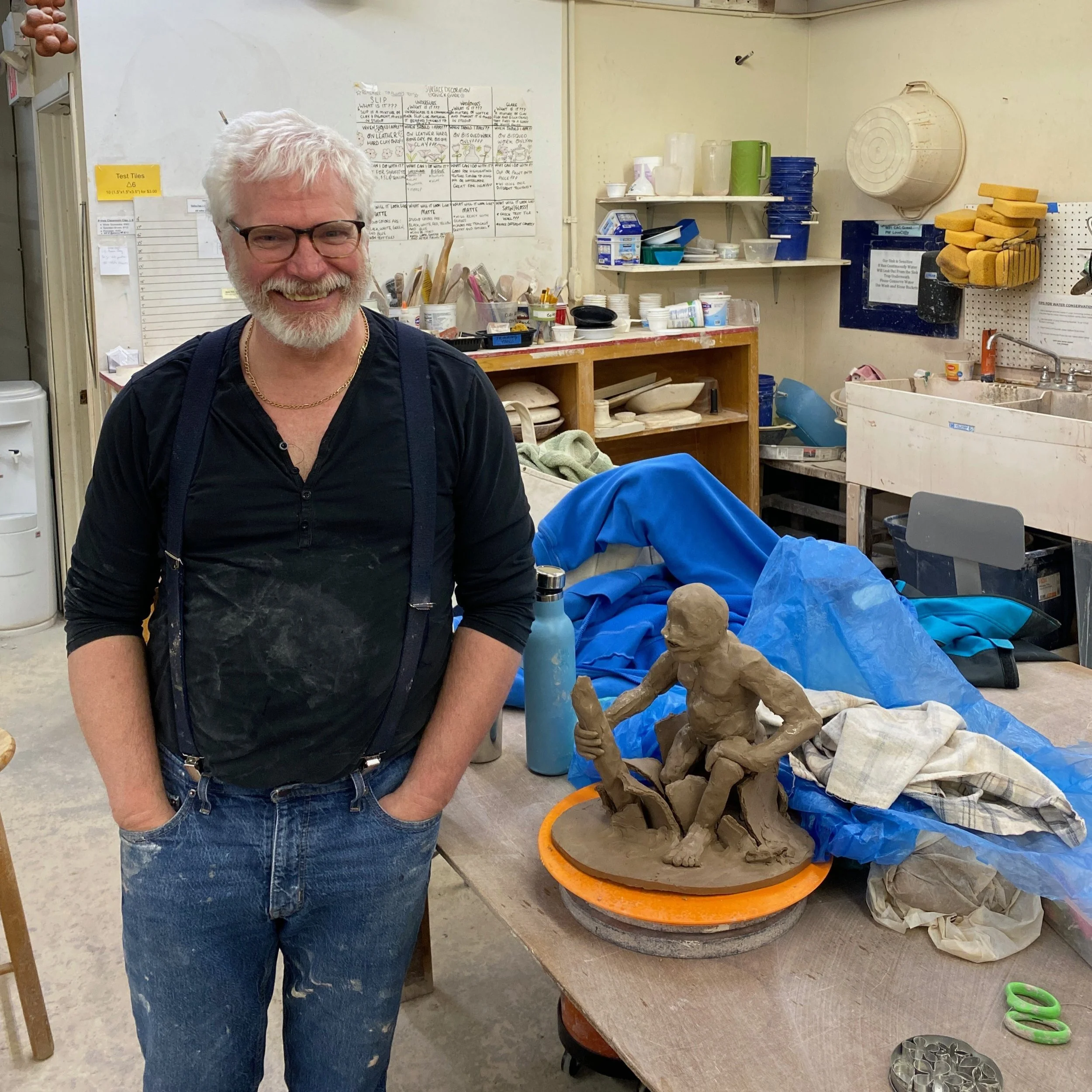 Jeff stands next to a work bench in the classroom with his hands in his pockets. On the bench is his sculpture of a seated humanoid figure, still in early stages.