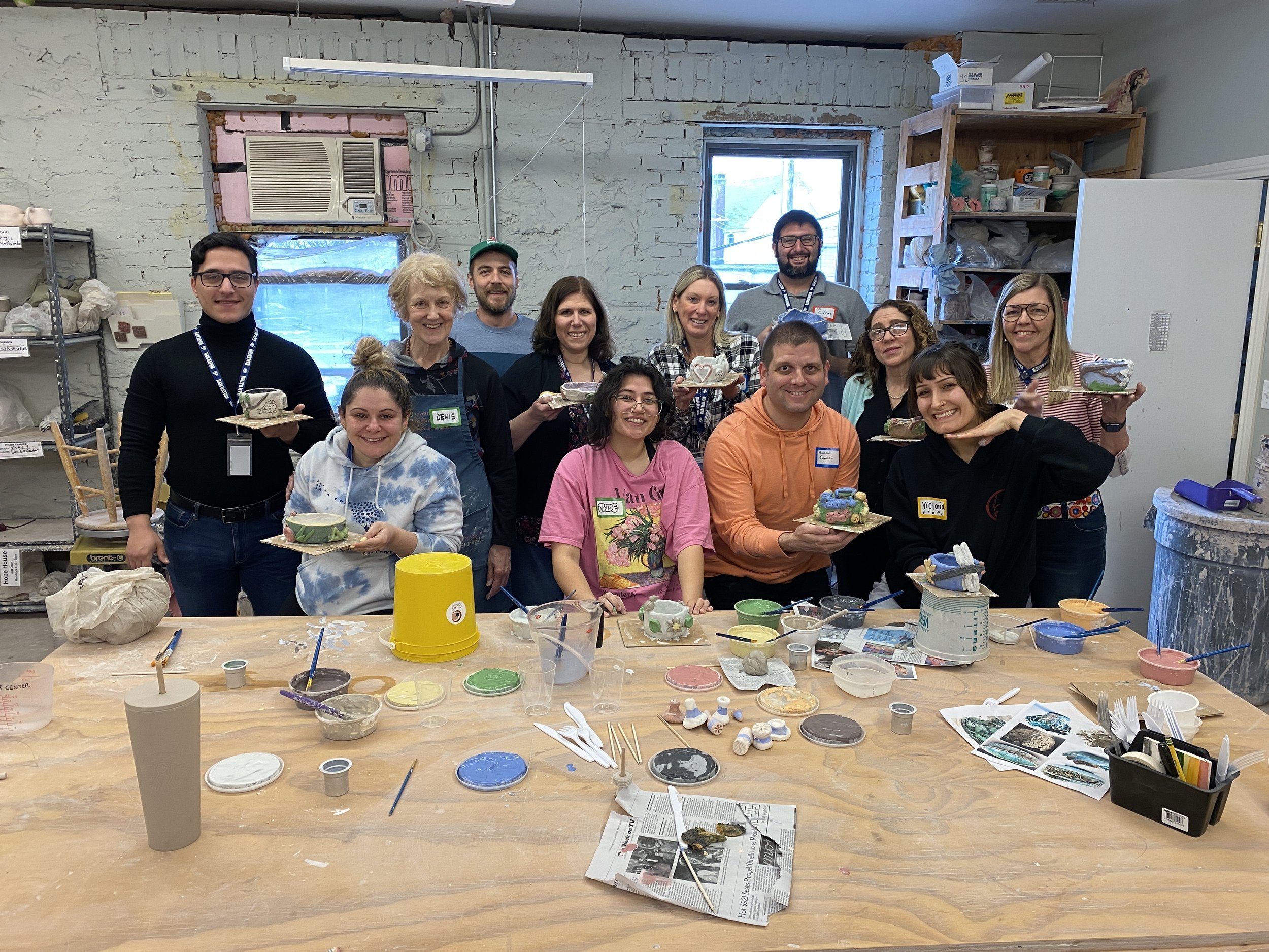 A group of Port Chester teachers hold up their clay pinch pots. In front of them is a wooden work bench strewn with open containers of bright underglaze and tools.