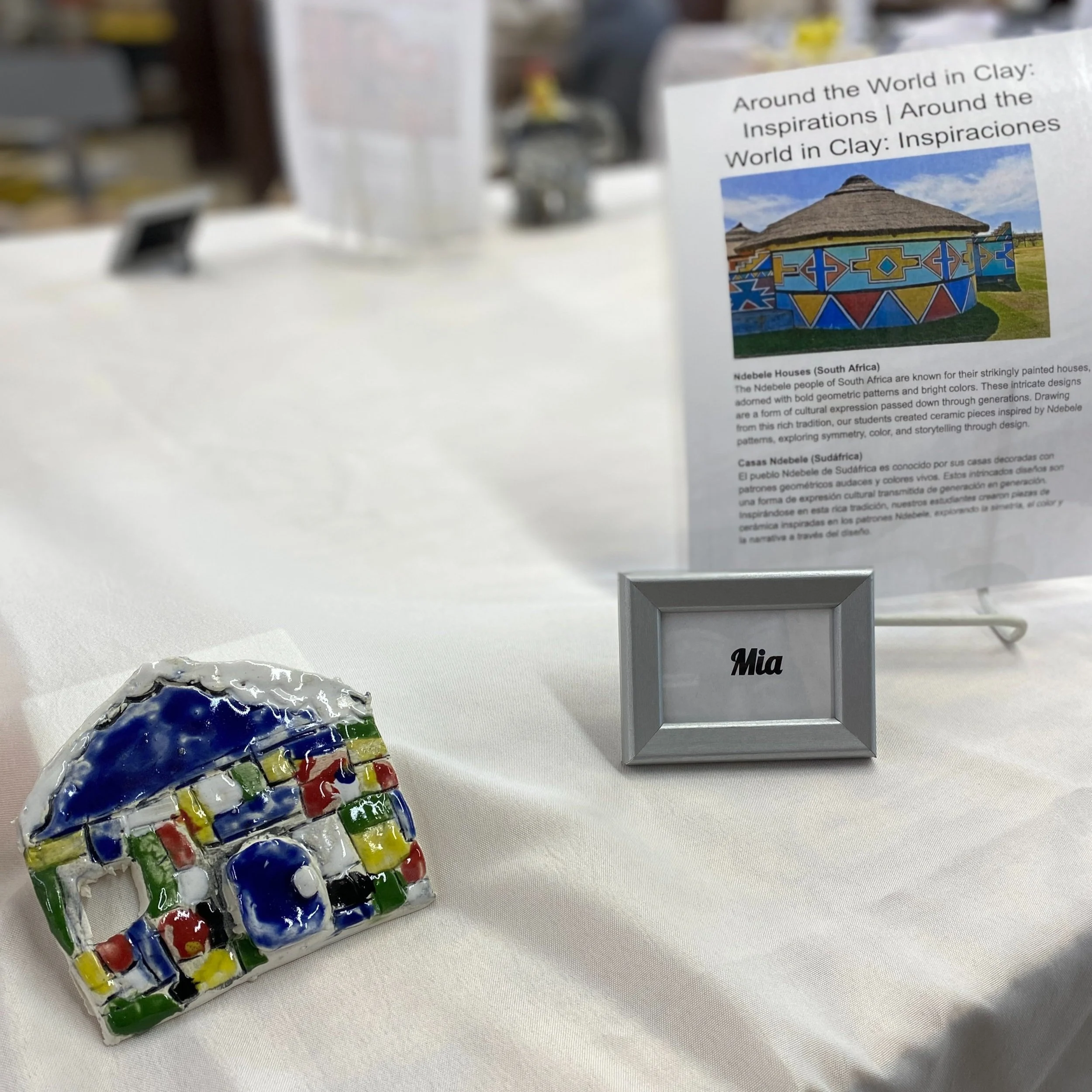 A colorful, flat, ceramic house lies on the table next to the name tag "Mia." Behind the name tag is a poster about the inspiration for the ceramic project in both English and Spanish.