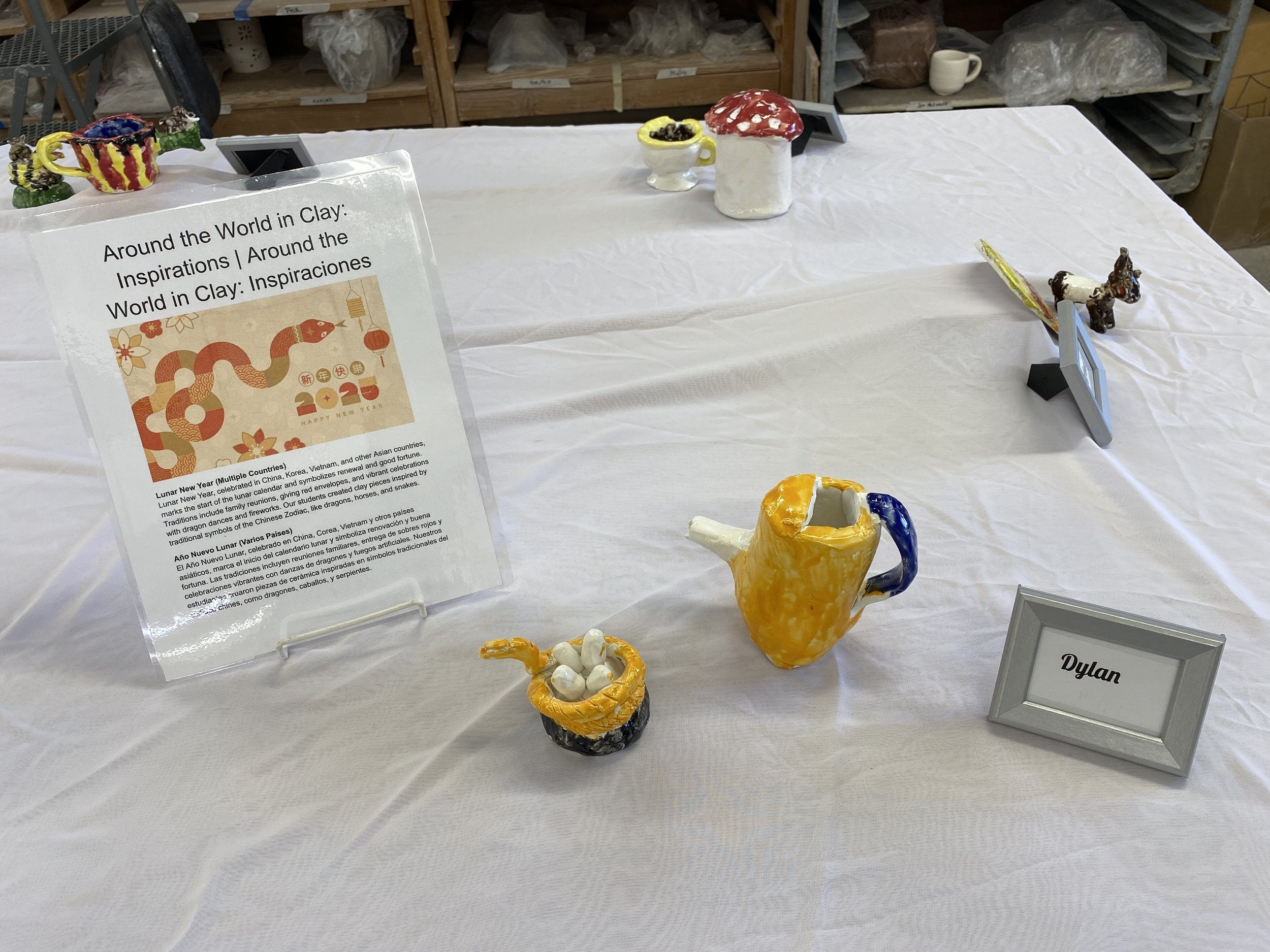 A small yellow teapot with a blue handle and other ceramic sculptures sit on the white tablecloth, along with a poster about the inspiration behind the projects in both English and Spanish.