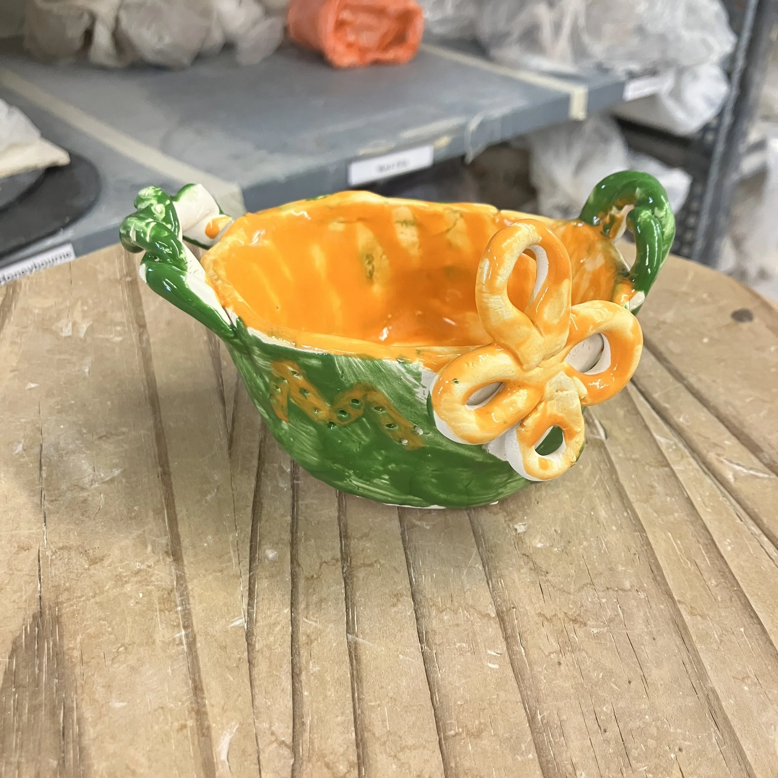 Small green pinchpot basket with a green exterior and orange interior. There is a decorative orange flower attached to the outside of the pot, and the basket handles are decoratively twisted.