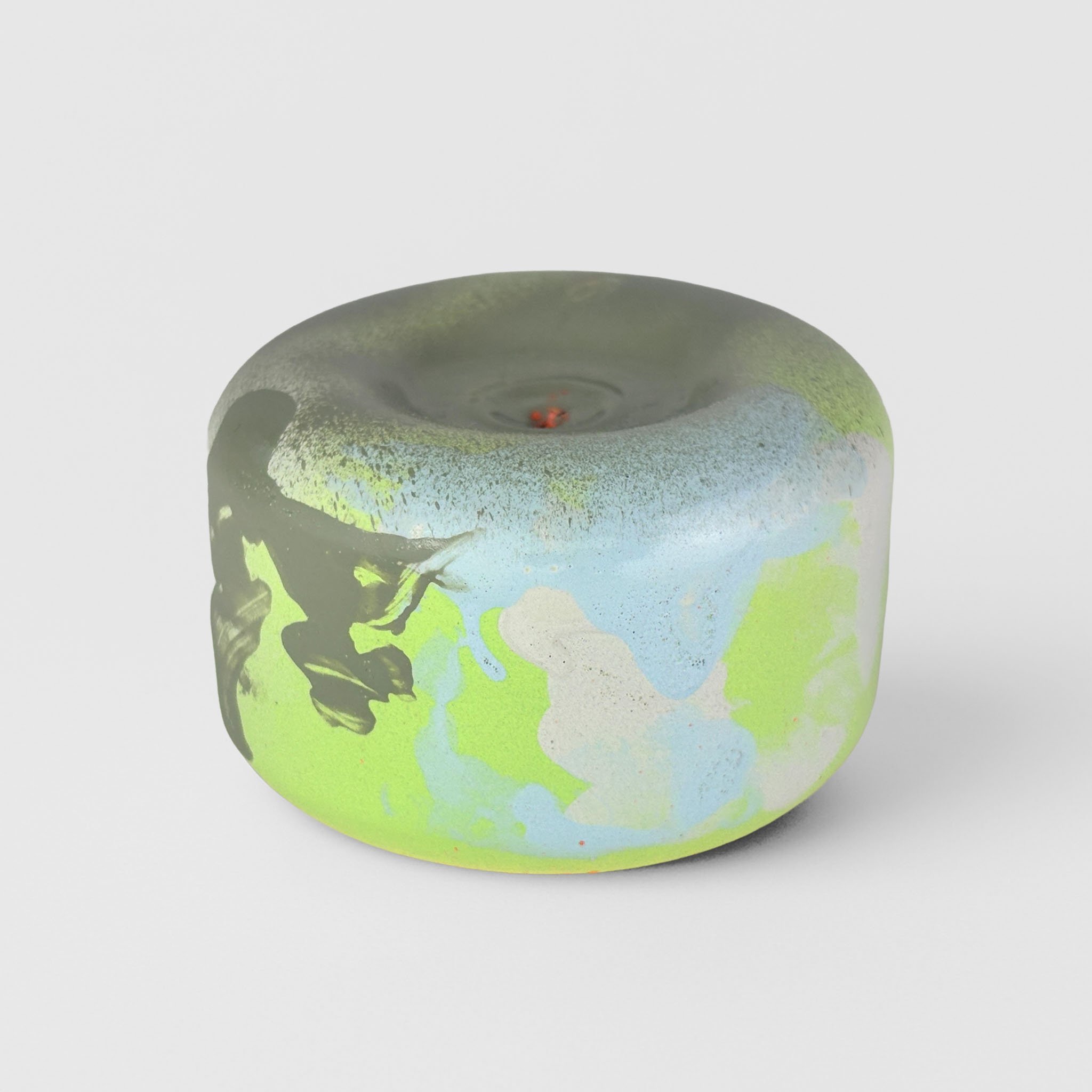 Close-up of a candle with abstract, colorful design in shades of green, blue, black, and white, placed on a white surface.