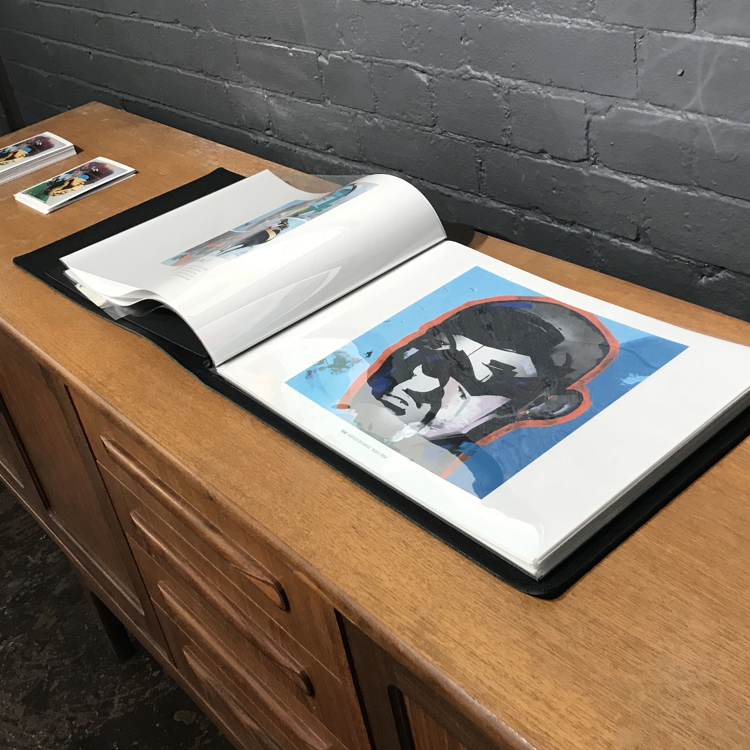Open art book displaying a street art style portrait of a person with sunglasses, surrounding a stylized orange outline, on a wooden surface against a gray brick wall.