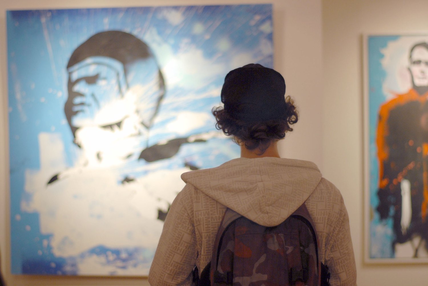 Person with dark curly hair wearing a beige hoodie and carrying a backpack, observing a large painting of a face with sunglasses, set in an art gallery.