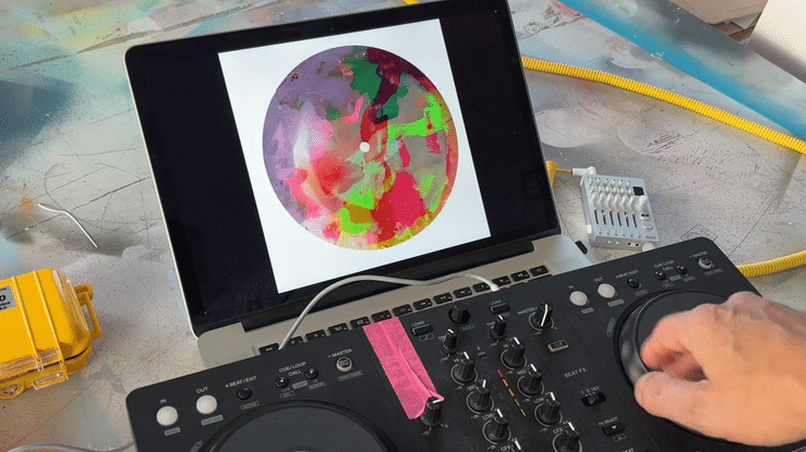 DJ mixing equipment connected to a laptop displaying a colorful abstract design, with a person's hand adjusting a control.
