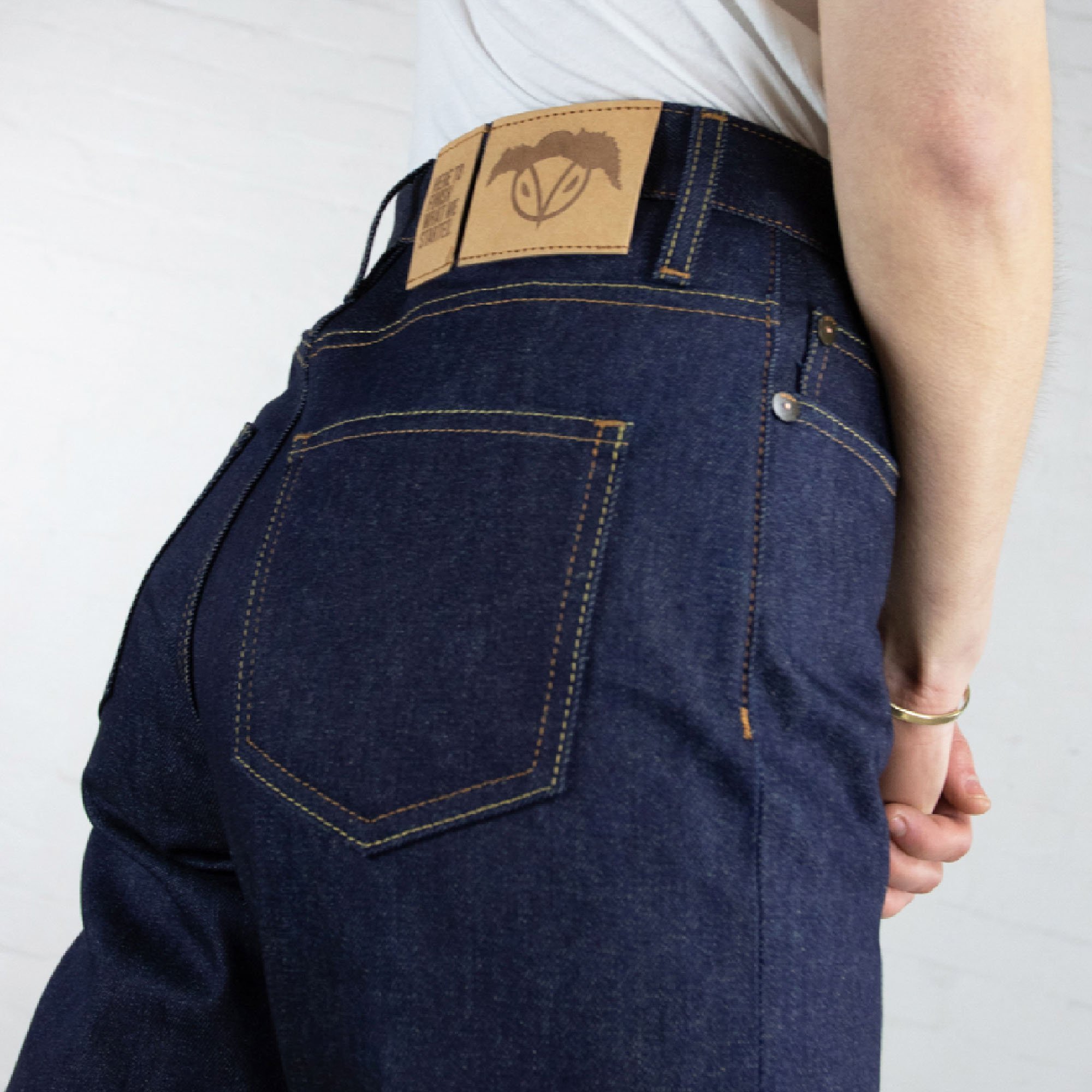 Close-up of person wearing dark blue jeans with leather patch on waistband and yellow stitching, standing against a white wall.