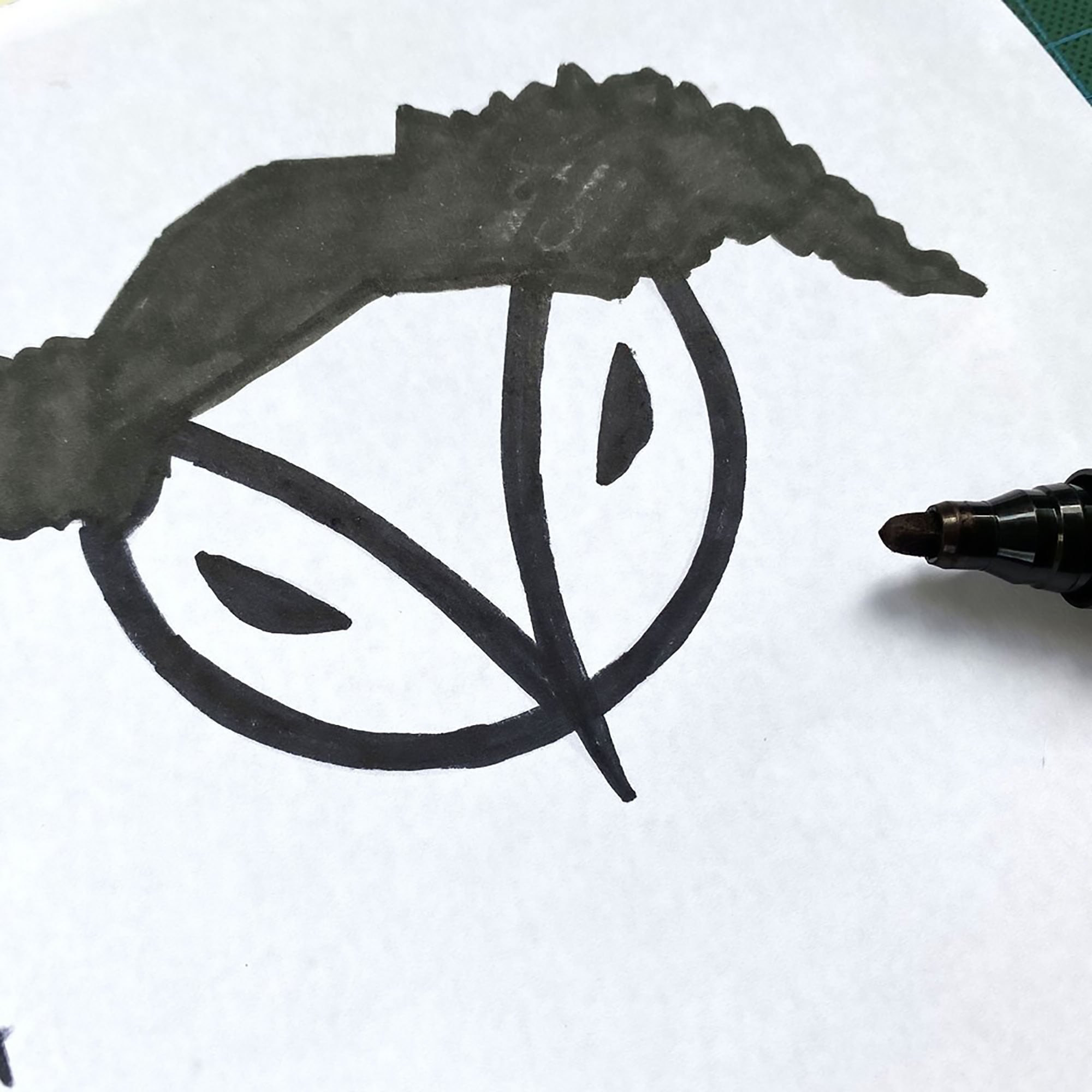 A black marker drawing of an abstract face with eyes and a nose on white paper, with the tip of the marker visible.