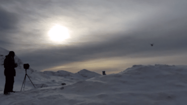 Photographer standing on snowy landscape capturing a sunset or sunrise with a camera on a tripod.